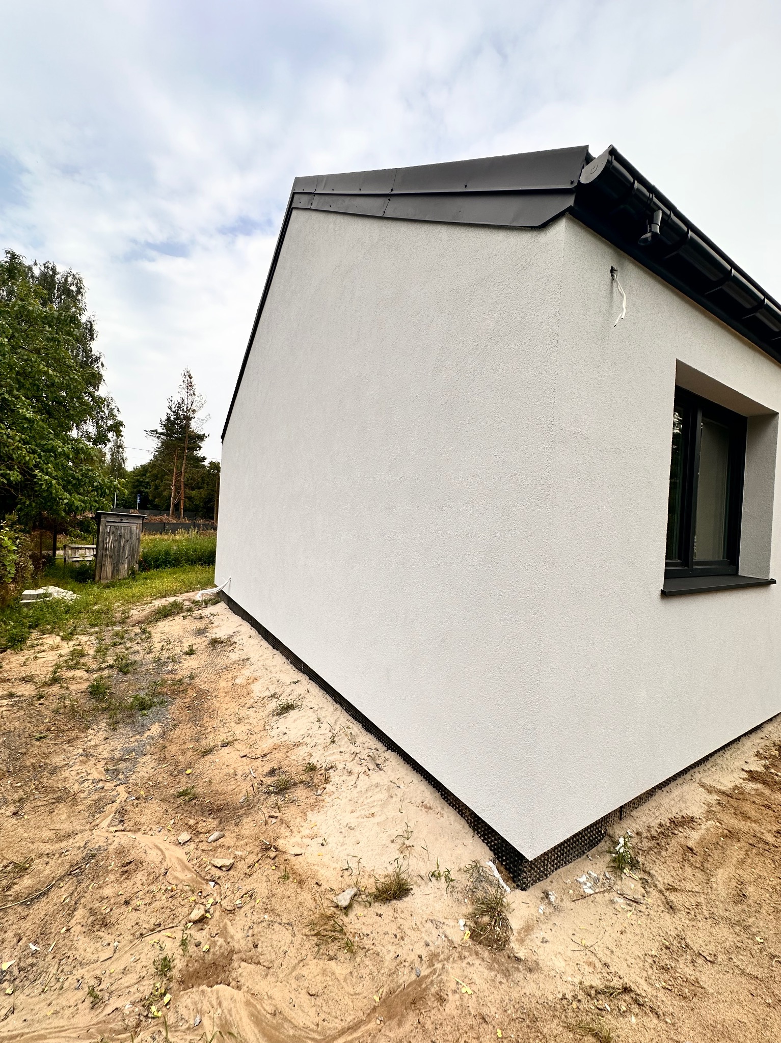 Nowo otynkowana ściana budynku z czarną stolarką okienną i wykończonym dachem, widoczna z poziomu gruntu z odsłoniętą ziemią wokół fundamentów.