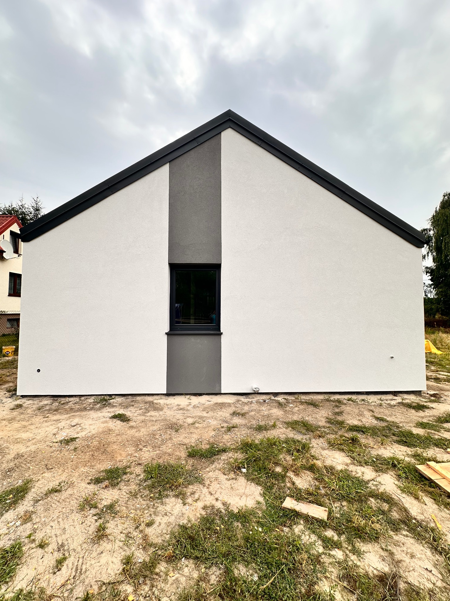 Nowoczesny dom jednorodzinny z białą elewacją i szarym akcentem wokół okna, widok z zewnątrz na tle pochmurnego nieba.