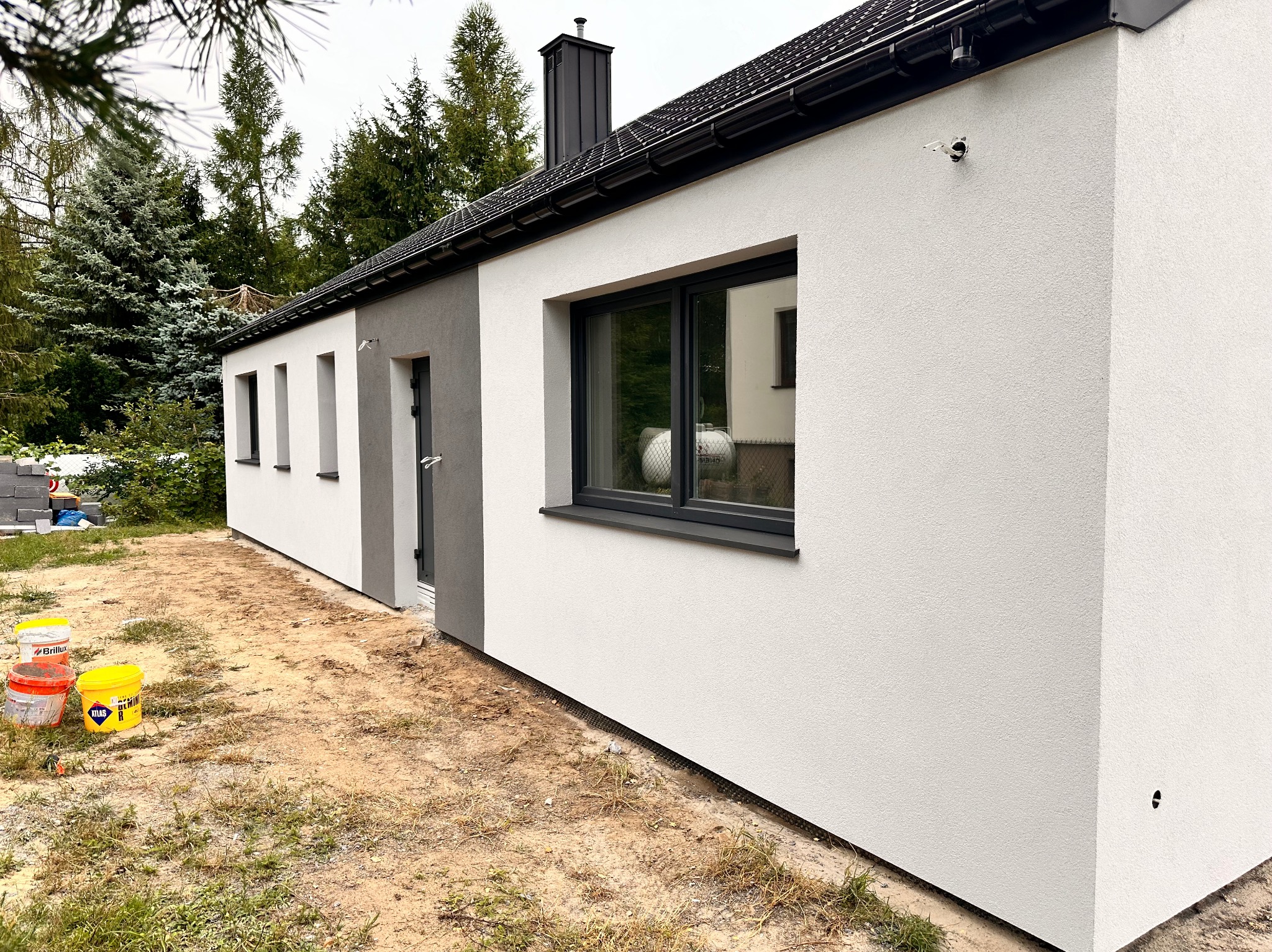 Świeżo otynkowana elewacja domu jednorodzinnego z widocznymi puszkami instalacyjnymi, czarną stolarką okienną i dachem z blachy na rąbek stojący; na pierwszym planie wiaderka po farbach elewacyjnych.