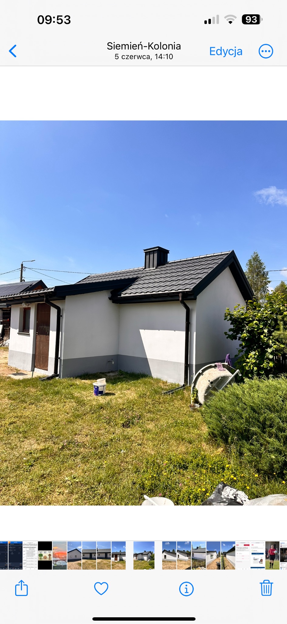 Biały budynek z czarnym dachem i rynnami, widoczne ślady świeżego malowania na elewacji i wiadro z farbą na trawniku.