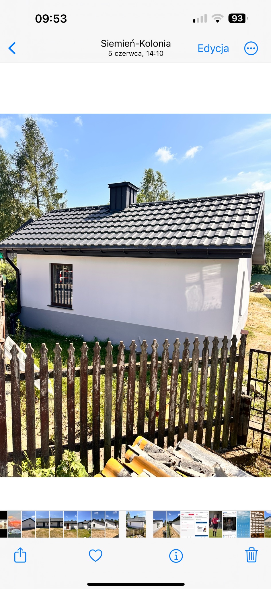 Widok z zewnątrz na otynkowany budynek z nowym dachem z blachodachówki w kolorze grafitowym, z widocznym kominem; w tle drzewa, a na pierwszym planie drewniany płot i stos starych dachówek.