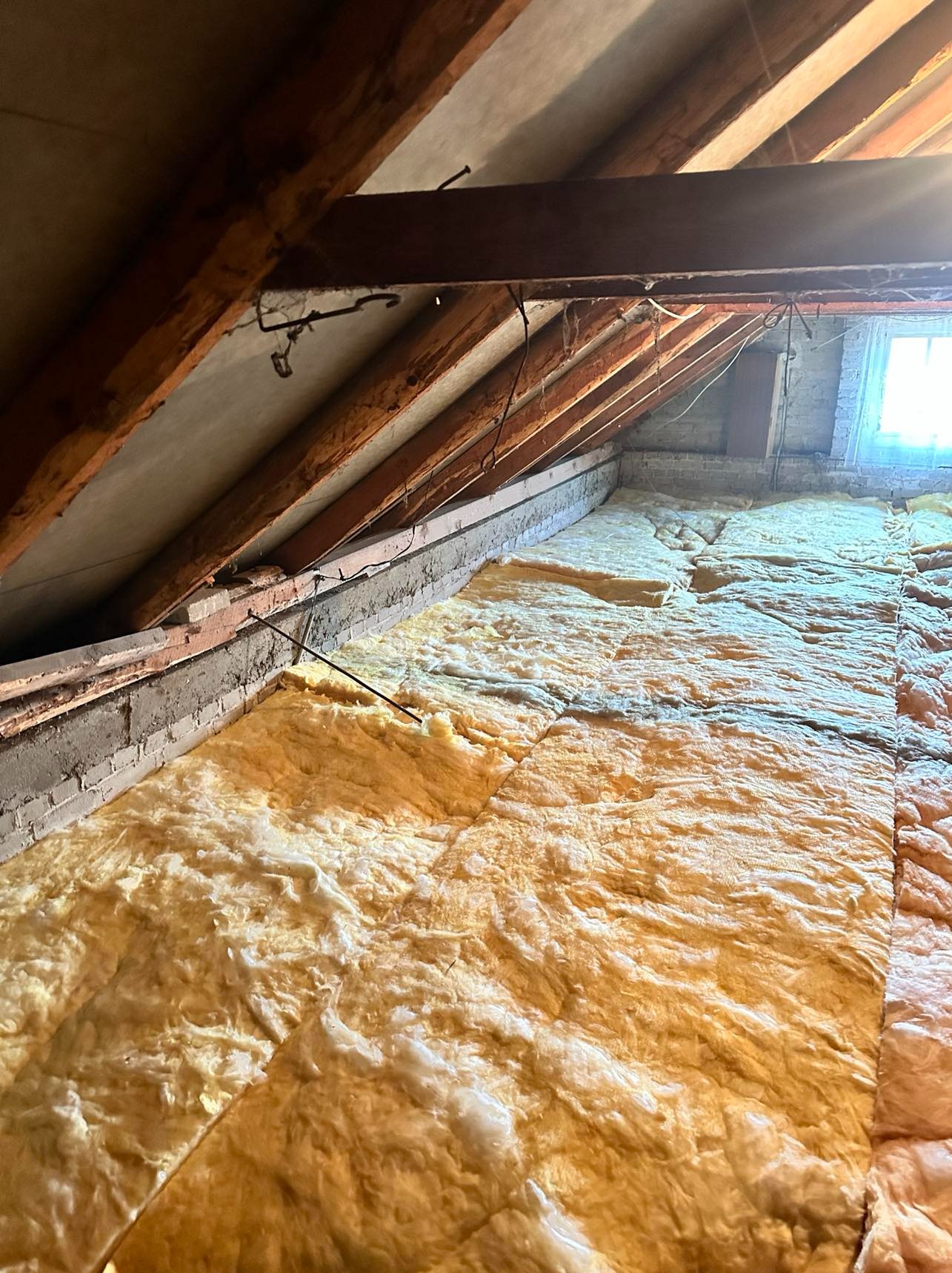 Poddasze z widoczną izolacją z wełny mineralnej, drewniane belki stropowe, mur z cegły i okno w tle.