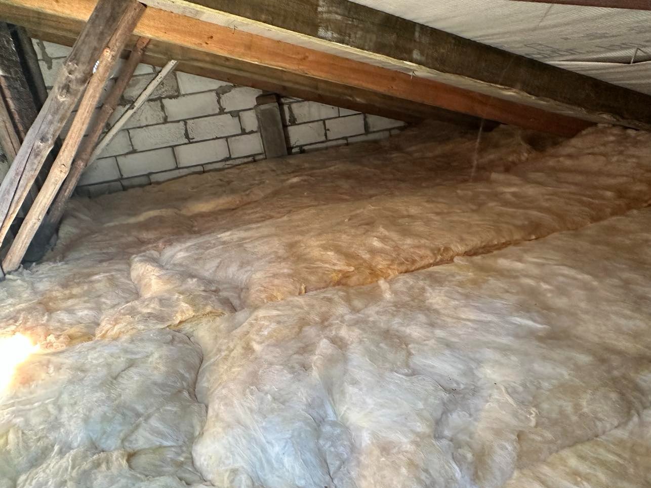 Poddasze z widoczną wełną mineralną pomiędzy drewnianymi belkami stropowymi i fragmentem ściany z bloczków betonowych.