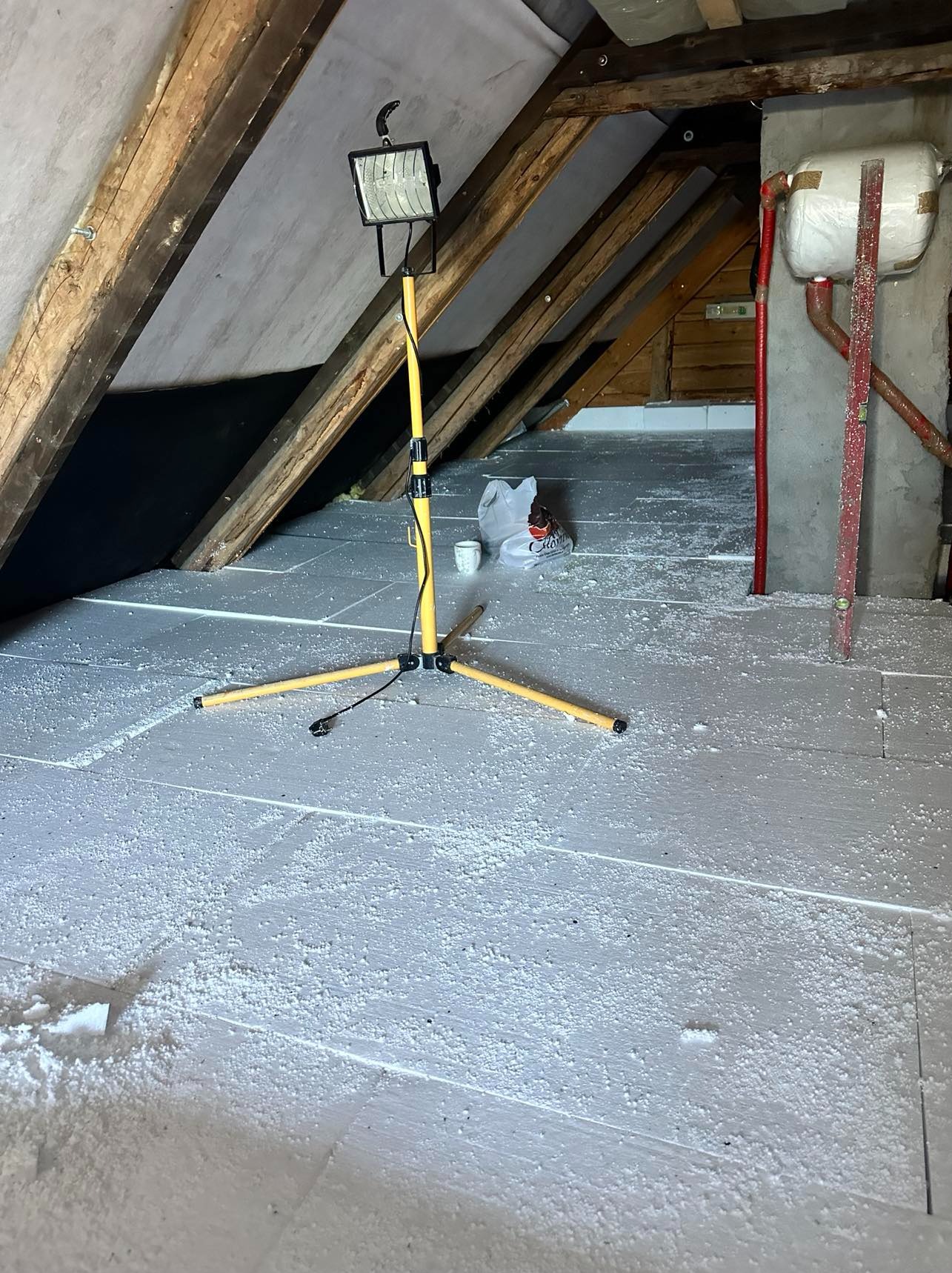 Poddasze w trakcie prac izolacyjnych z widoczną konstrukcją drewnianą, oświetleniem roboczym na statywie, rozłożonymi płytami izolacyjnymi i drobnymi resztkami materiału izolacyjnego na podłodze...