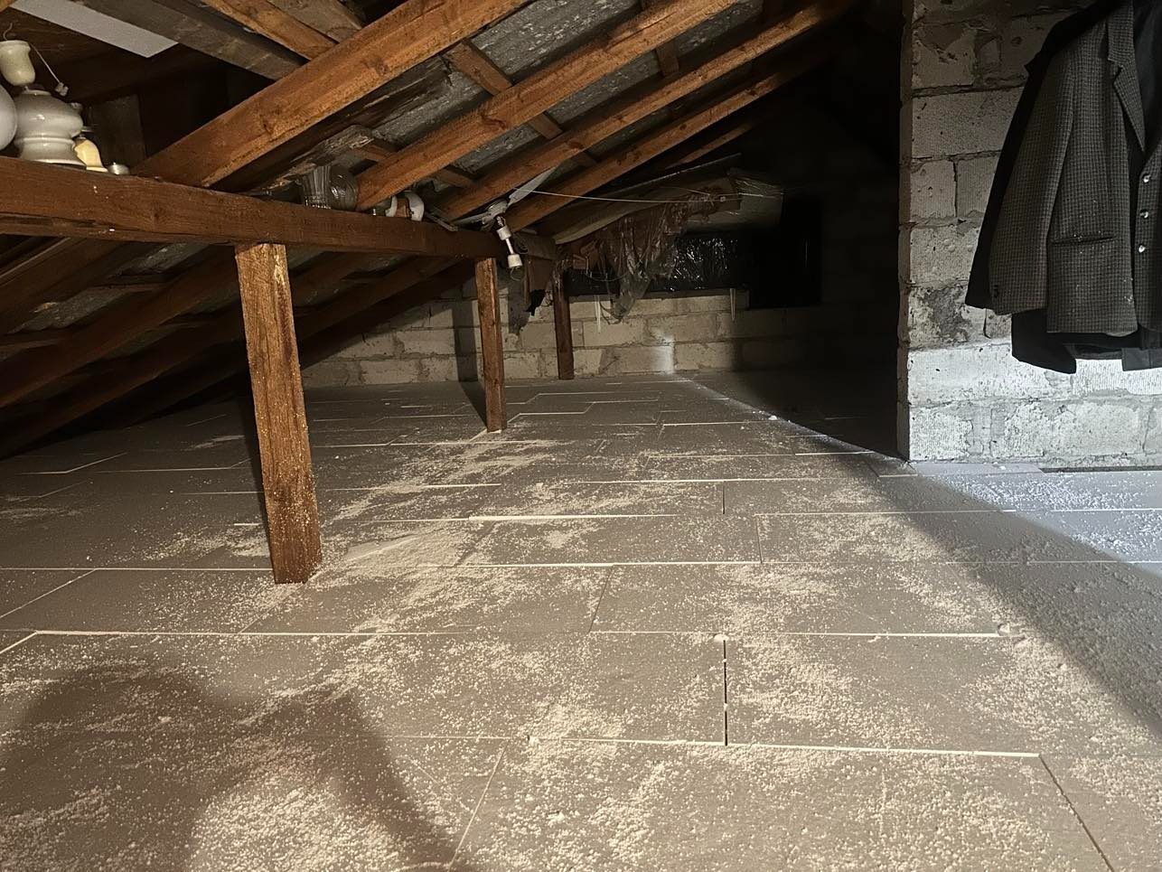 Poddasze z drewnianą konstrukcją dachu, wyłożone szarymi płytami podłogowymi pokrytymi białym pyłem, z widocznym fragmentem murowanej ściany i wiszącą marynarką.