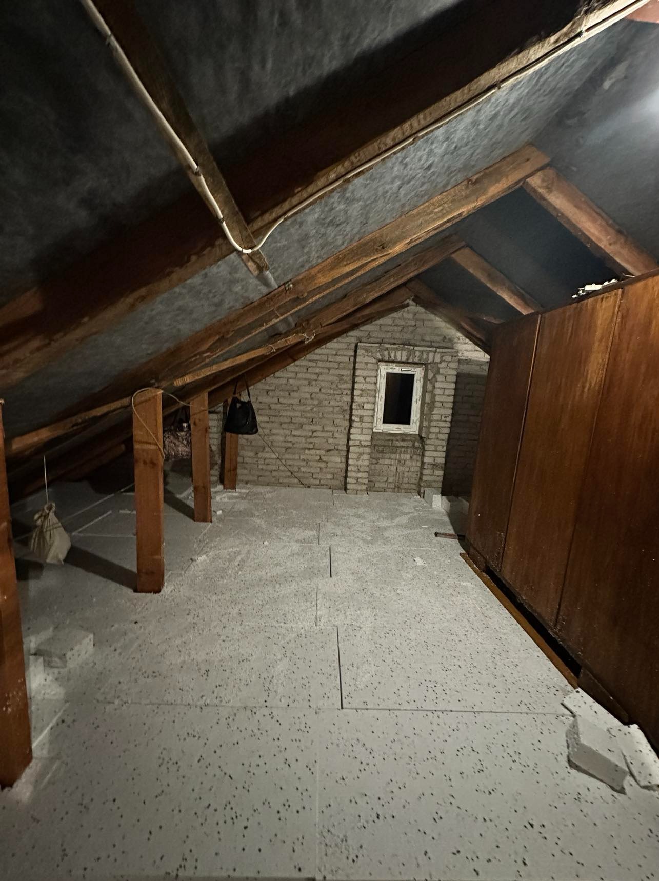 Poddasze z widoczną konstrukcją drewnianą dachu, ocieplone płytami styropianowymi, z fragmentem murowanej ściany z oknem i starą szafą.