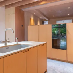 Nowoczesna kuchnia z jasnego drewna z wbudowanym piekarnikiem, chromowaną baterią przy zlewie i widoczną strukturą drewnianego stropu