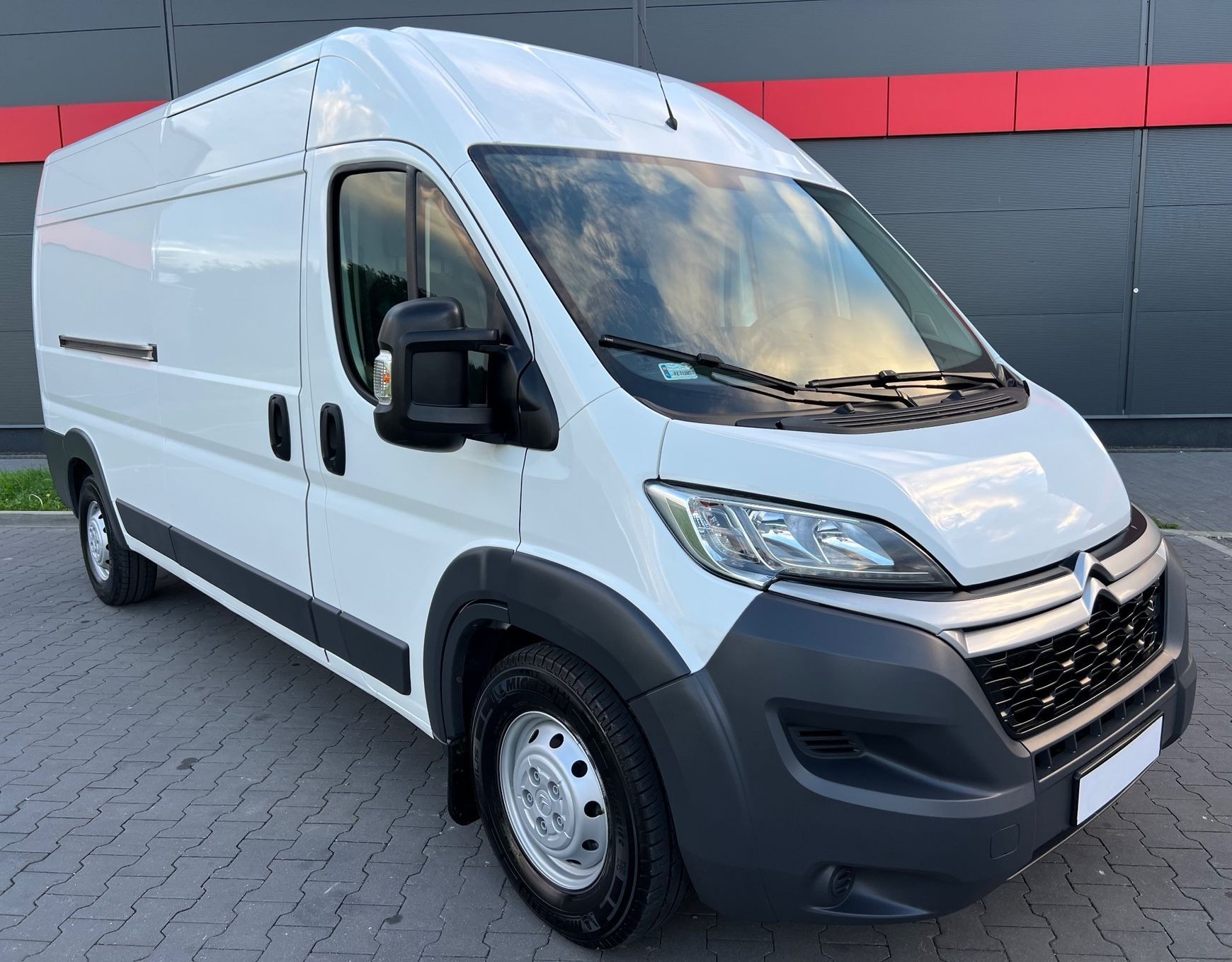 Biały samochód dostawczy marki Citroen z widocznym odbiciem chmur na przedniej szybie, zaparkowany na szarym bruku na tle szaro-czerwonej ściany budynku.