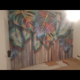 Ekodom - Ściana z fototapetą w motywie tropikalnych liści na pionowych panelach imitujących drewno, częściowo zakryta kartonem na podłodze w trakcie prac.