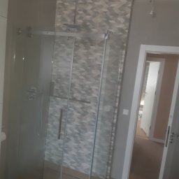 Ekodom - Szklana kabina prysznicowa z deszczownicą w łazience wykończonej płytkami w geometryczny wzór, widok na otwarte drzwi do kolejnego pomieszczenia.