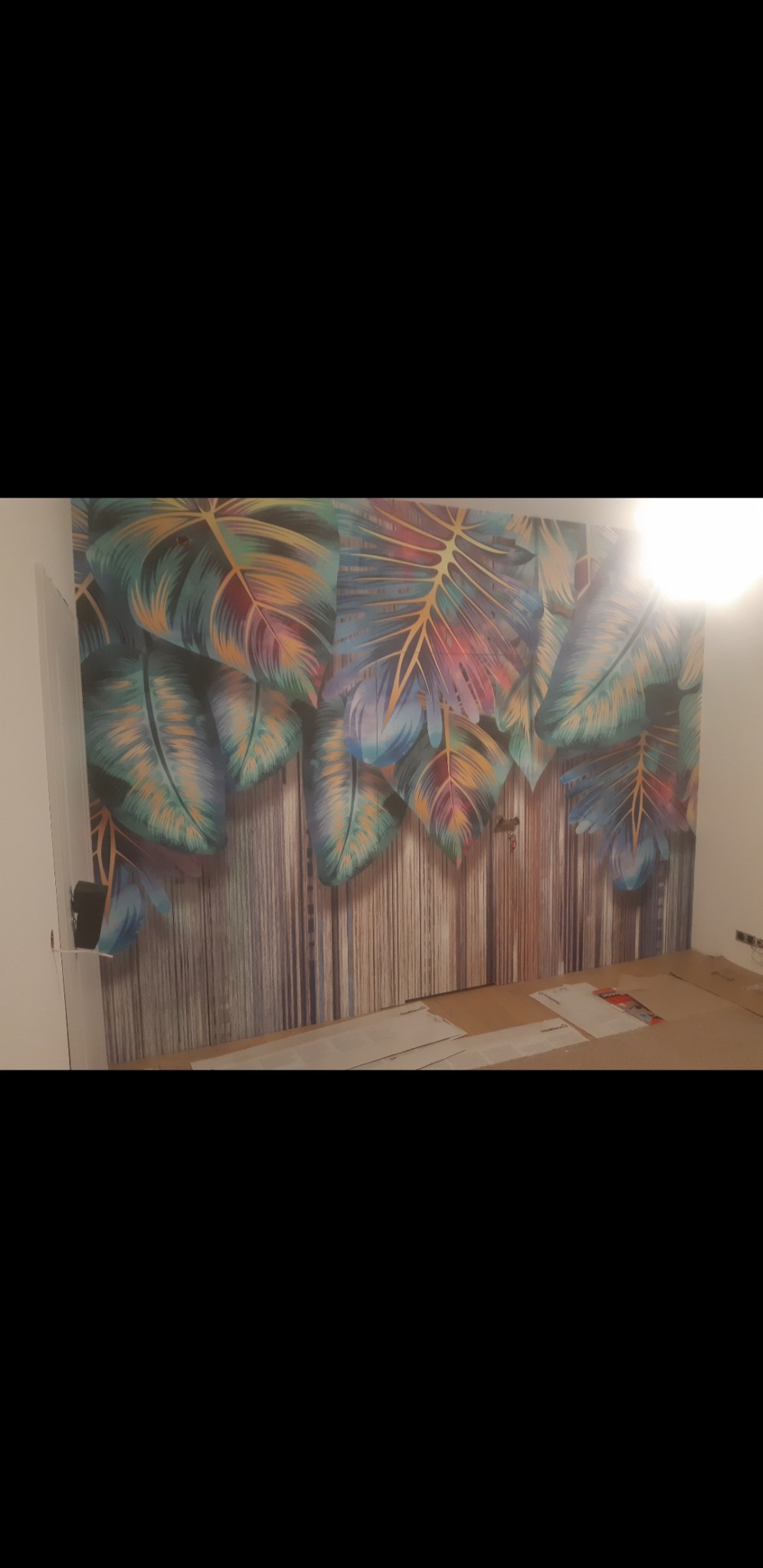 Ściana z fototapetą w motywie tropikalnych liści na pionowych panelach imitujących drewno, częściowo zakryta kartonem na podłodze w trakcie prac.