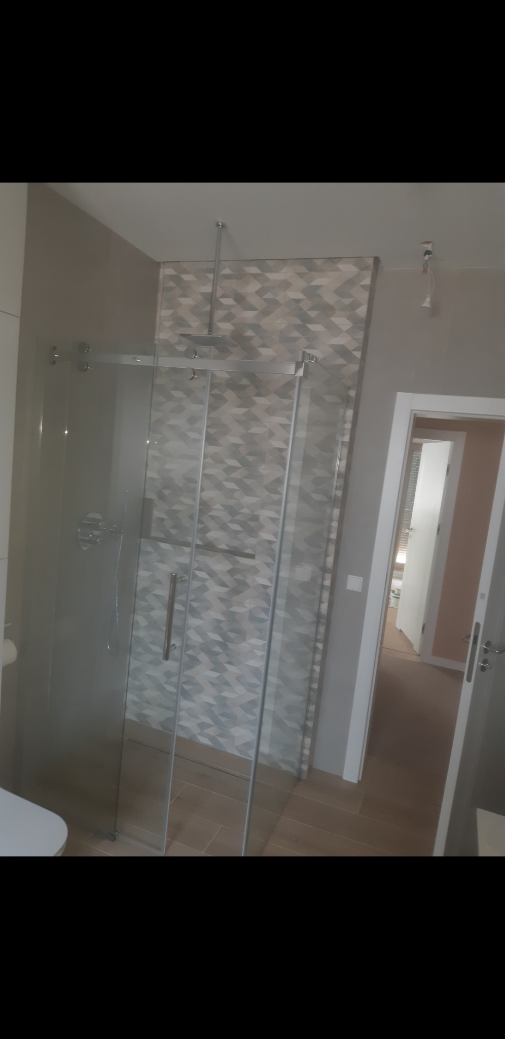 Szklana kabina prysznicowa z deszczownicą w łazience wykończonej płytkami w geometryczny wzór, widok na otwarte drzwi do kolejnego pomieszczenia.