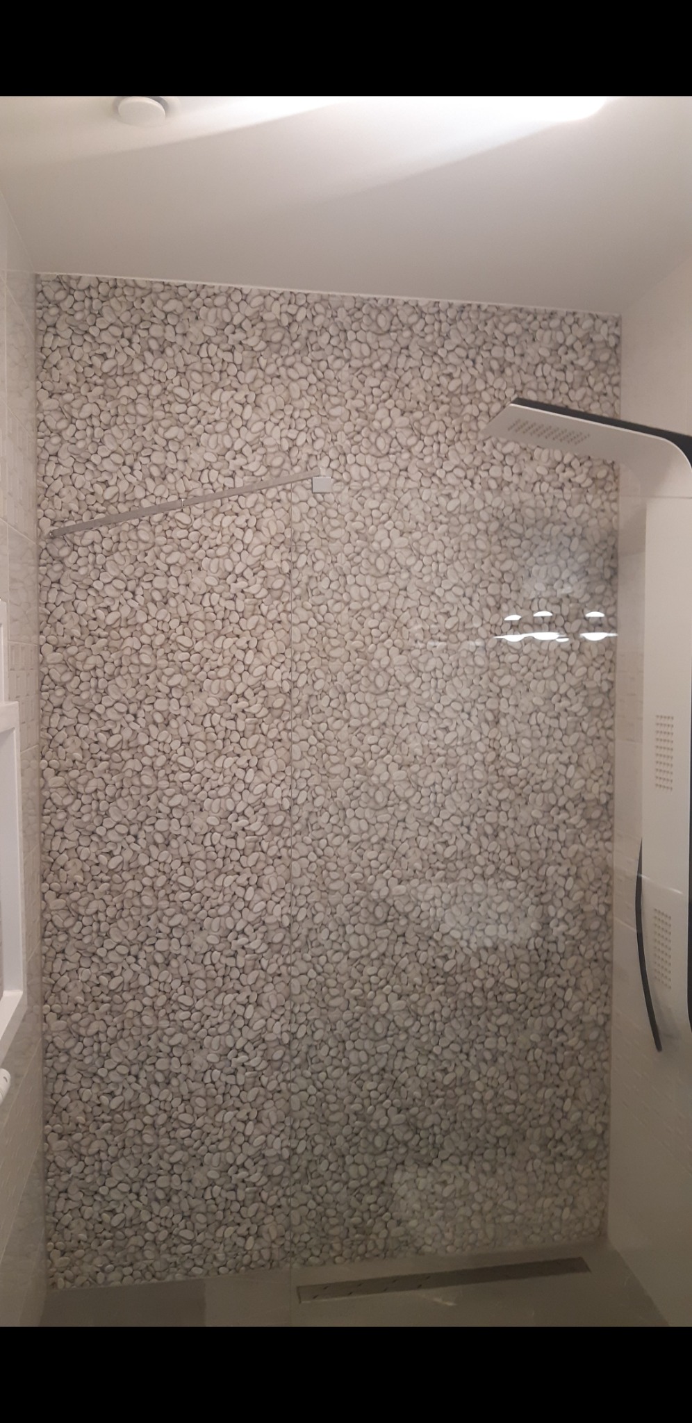 Kabina prysznicowa z wyłożoną drobnymi, beżowymi kamieniami ścianą, szklana szyba z widocznym odpływem liniowym w podłodze i nowoczesną deszczownicą.