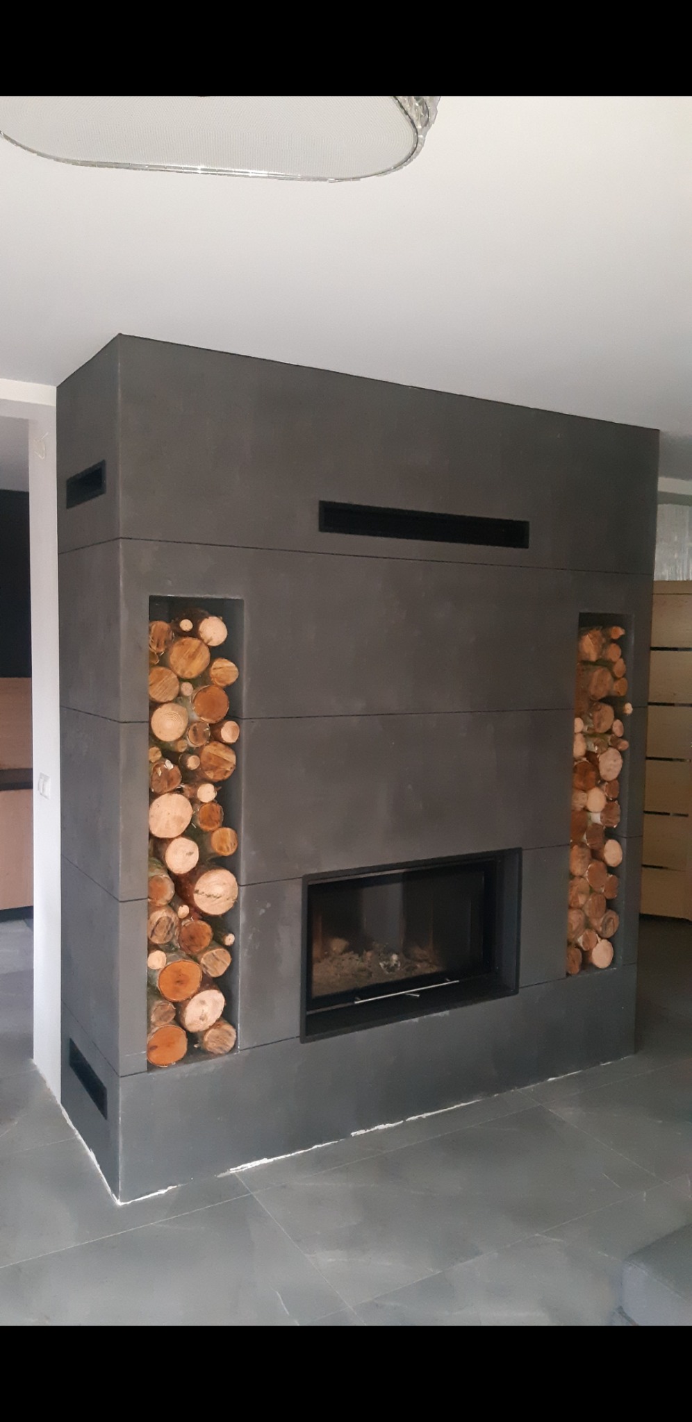 Nowoczesny kominek z obudową z szarego betonu architektonicznego, z wnękami wypełnionymi drewnem opałowym po bokach i czarnym wkładem kominkowym.