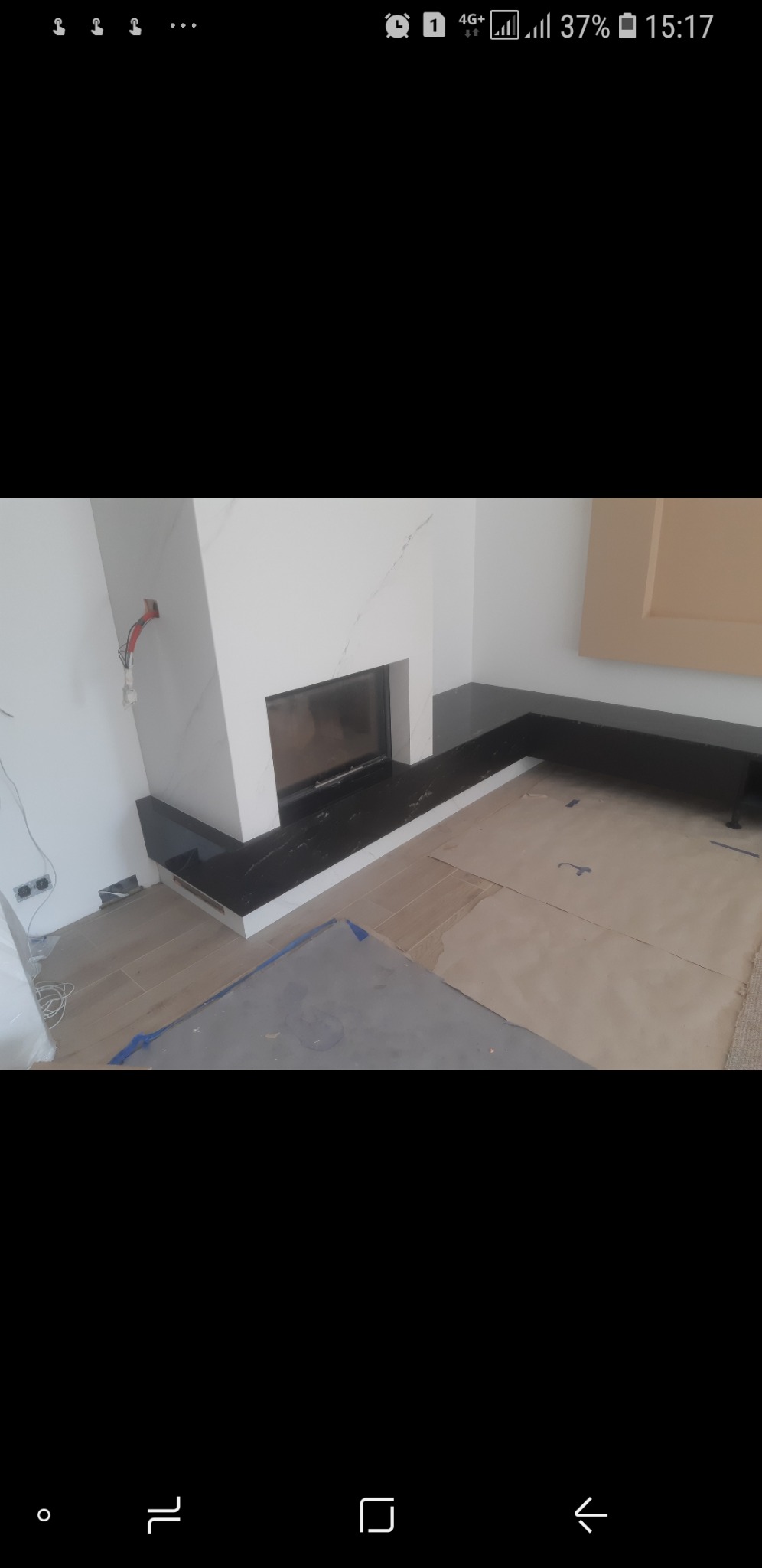 Nowoczesny kominek z białego marmuru z czarnym blatem, częściowo zabezpieczony folią malarską, widoczne gniazdka elektryczne i elementy instalacji.