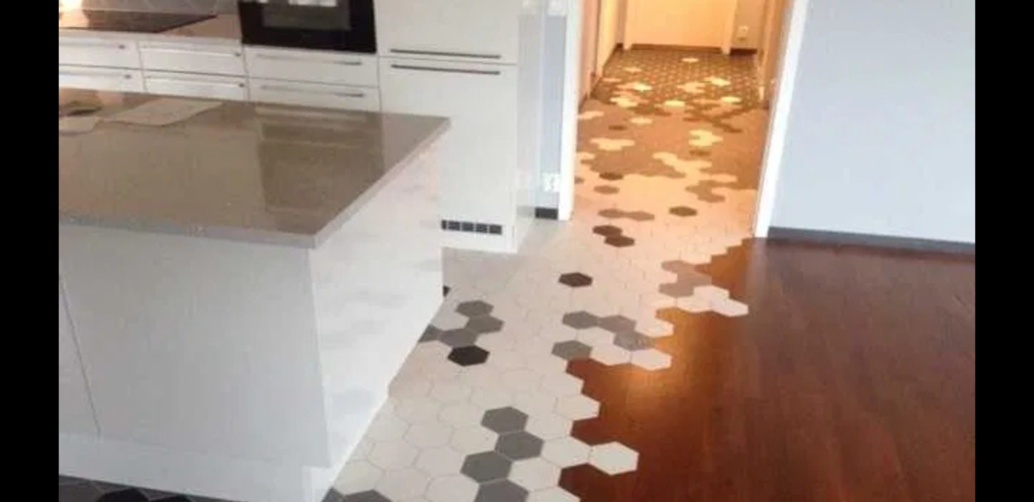 Kuchenna podłoga przechodzi w parkiet dzięki heksagonalnym płytkom w odcieniach bieli, szarości i czerni, tworząc płynne przejście kolorystyczne i materiałowe.