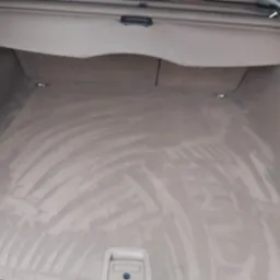 Jasnobeżowa tapicerka bagażnika samochodu po czyszczeniu, widoczne delikatne smugi po odkurzaczu lub szczotce, plastikowa osłona w g&oacute;rnej części.