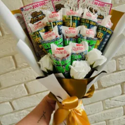 Bukiet z białych róż, słodyczy Raffaello i zdrapek loteryjnych 'Moc Szczęścia' i 'Super Szczęście', owinięty w biały i złoty papier, trzymany w dłoni z tatuażem na tle białej ceglanej ściany.