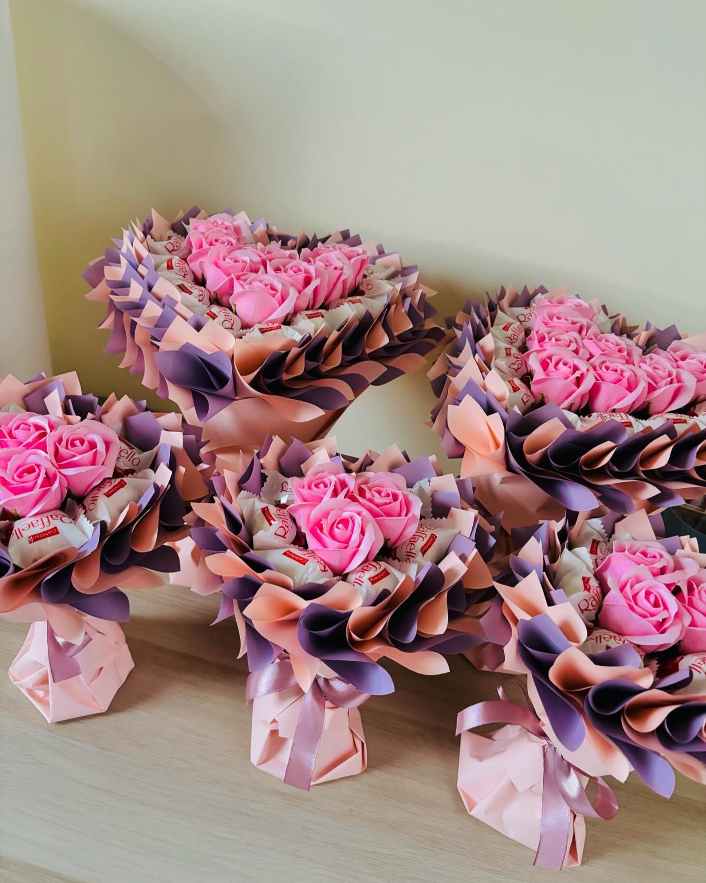 Kilka małych bukietów z różowych róż mydlanych i słodyczy Raffaello, zapakowanych w ozdobny papier w odcieniach fioletu i różu, ustawionych na drewnianym stole.
