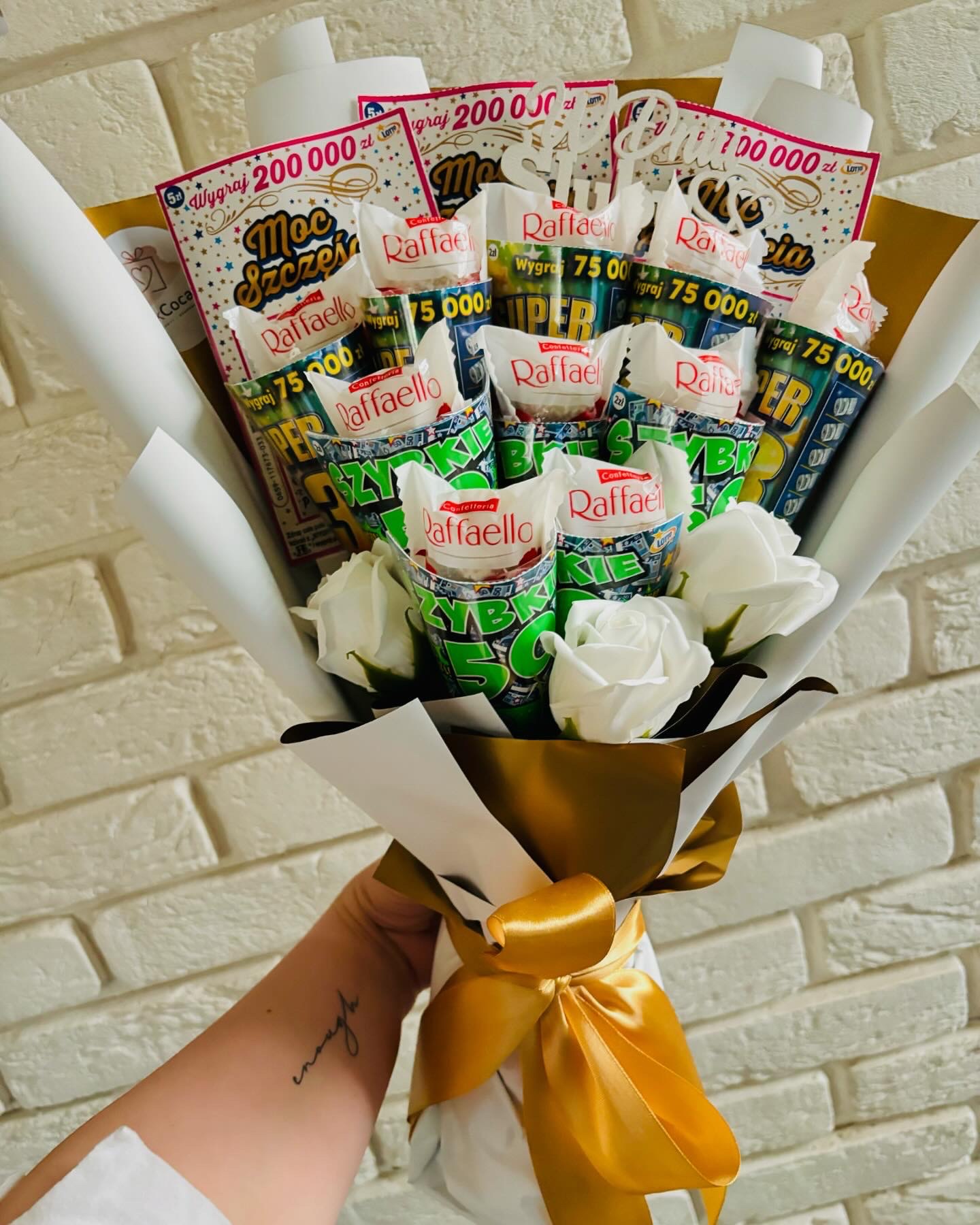 Bukiet z białych róż, słodyczy Raffaello i zdrapek loteryjnych 'Moc Szczęścia' i 'Super Szczęście', owinięty w biały i złoty papier, trzymany w dłoni z tatuażem na tle białej ceglanej ściany.