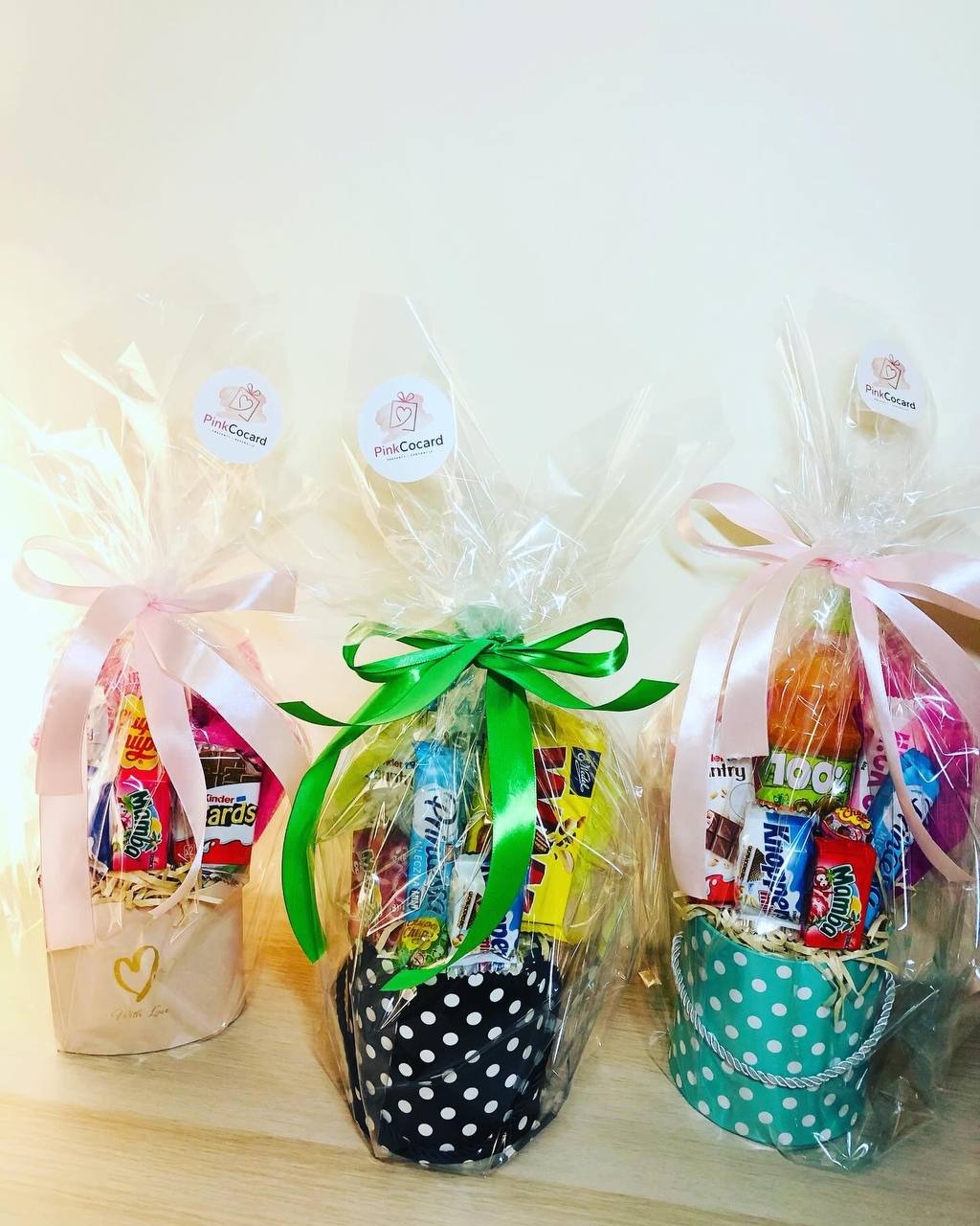 Trzy ozdobne, celofanowe paczki ze słodyczami, każda w innym, wzorzystym pojemniku: beżowym z sercem, czarnym w białe kropki i turkusowym w białe kropki, zawiązane odpowiednio różową, zieloną...