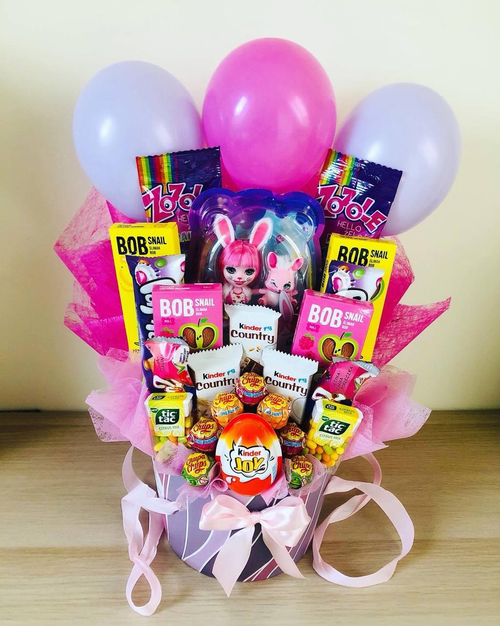 Różowy kosz z zabawkami i słodyczami, ozdobiony pastelowymi balonami i wstążkami, prezentujący różnorodność smakołyków i zabawek w estetycznej kompozycji.