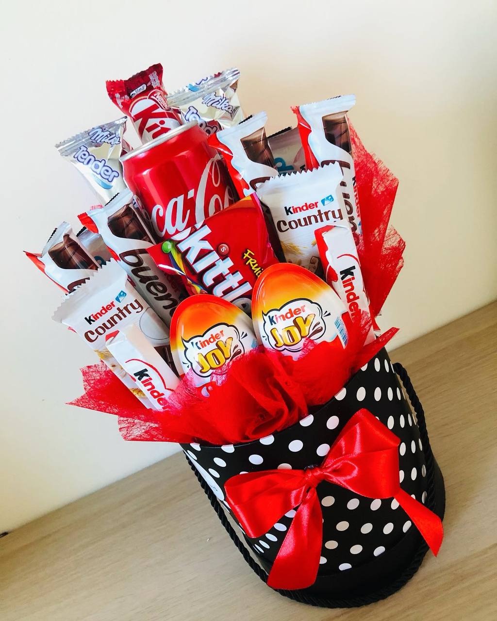 Słodki upominek w czarno-białym pudełku w kropki, wypełniony czerwonym tiulem, zawierający Coca-Colę, Kinder Joy, Kinder Country, KitKat oraz inne słodycze, przewiązany czerwoną wstążką