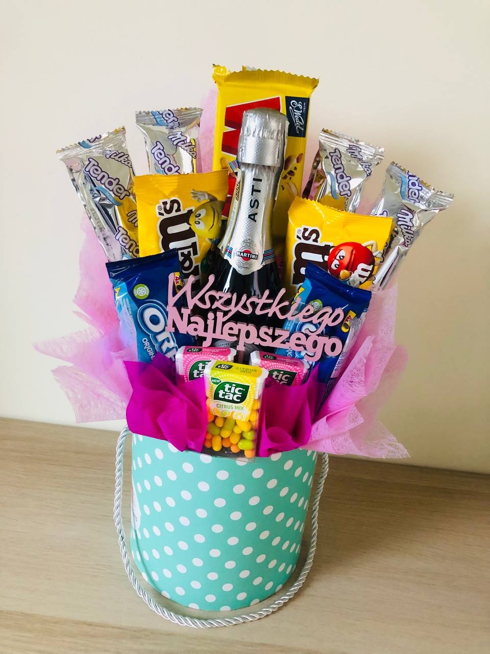 Słodki prezent w okrągłym, turkusowym pudełku w białe kropki, wypełniony chrupkami, batonami, cukierkami, mini butelką Martini i różową dekoracyjną bibułą, z napisem 'Wszystkiego Najlepszego'...