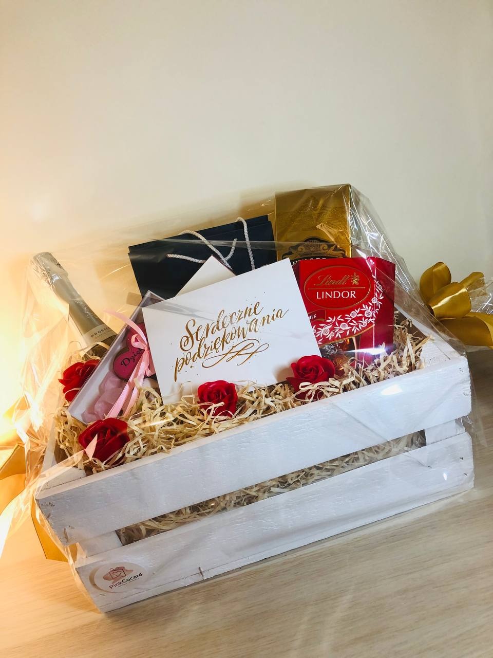 Biała drewniana skrzynka wypełniona siankiem, zawierająca butelkę Prosecco, czekoladki Lindor, małe torebki prezentowe, kartkę z napisem 'Serdeczne podziękowania' oraz dekoracyjne czerwone róże...