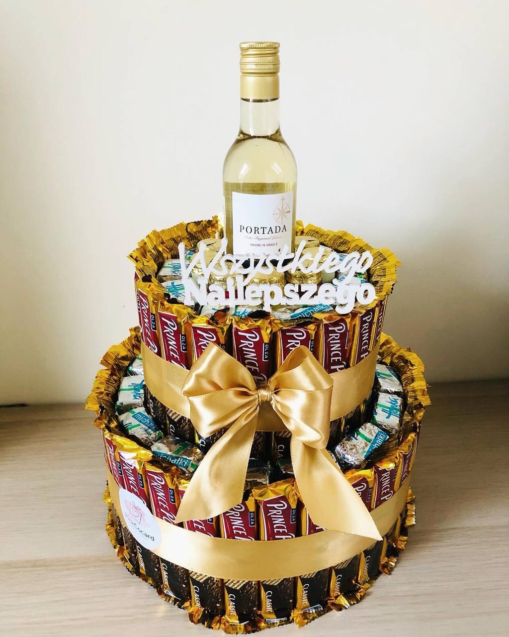 Dwupoziomowy tort z czekoladowych batoników Prince Polo, ozdobiony złotą wstążką z kokardą i butelką białego wina na górze, dekoracja z napisem 'Wszystkiego Najlepszego'.