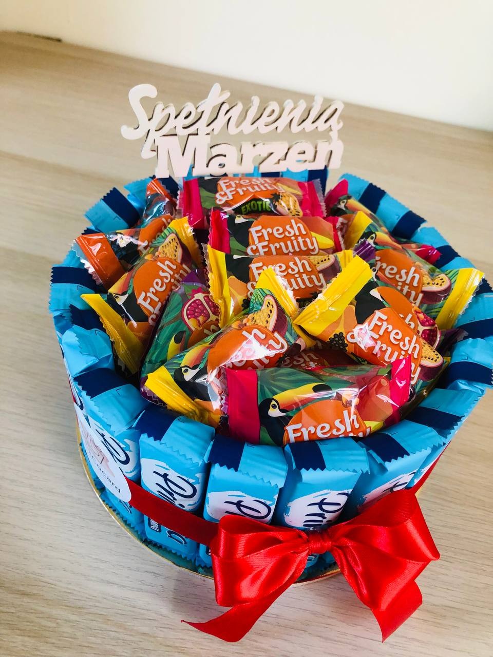 Dekoracyjny kosz wypełniony słodyczami Fresh Fruity, otoczony niebieskimi batonikami i przewiązany czerwoną wstążką, z napisem 'Spełnienia Marzeń' na górze.