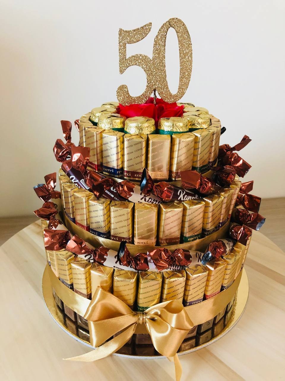 Trzypiętrowy tort z czekoladek Merci i cukierków Roshen, udekorowany złotą cyfrą 50 i czerwoną różą na szczycie, owinięty złotą wstążką z kokardą.