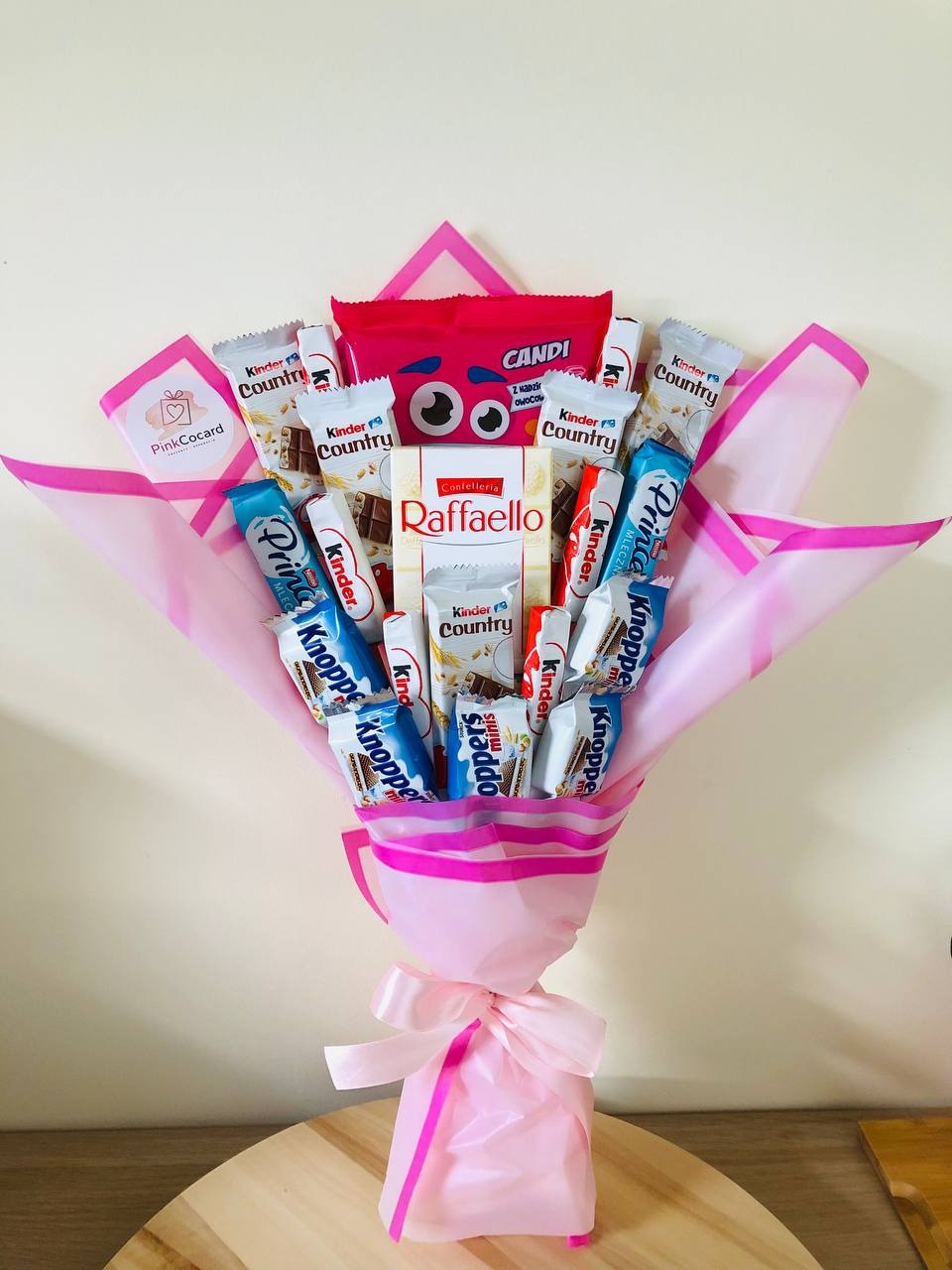 Słodki bukiet z batoników Kinder Country, Prince Polo i Raffaello, owinięty w różowy papier i ozdobiony różową kokardą, na drewnianym stole.