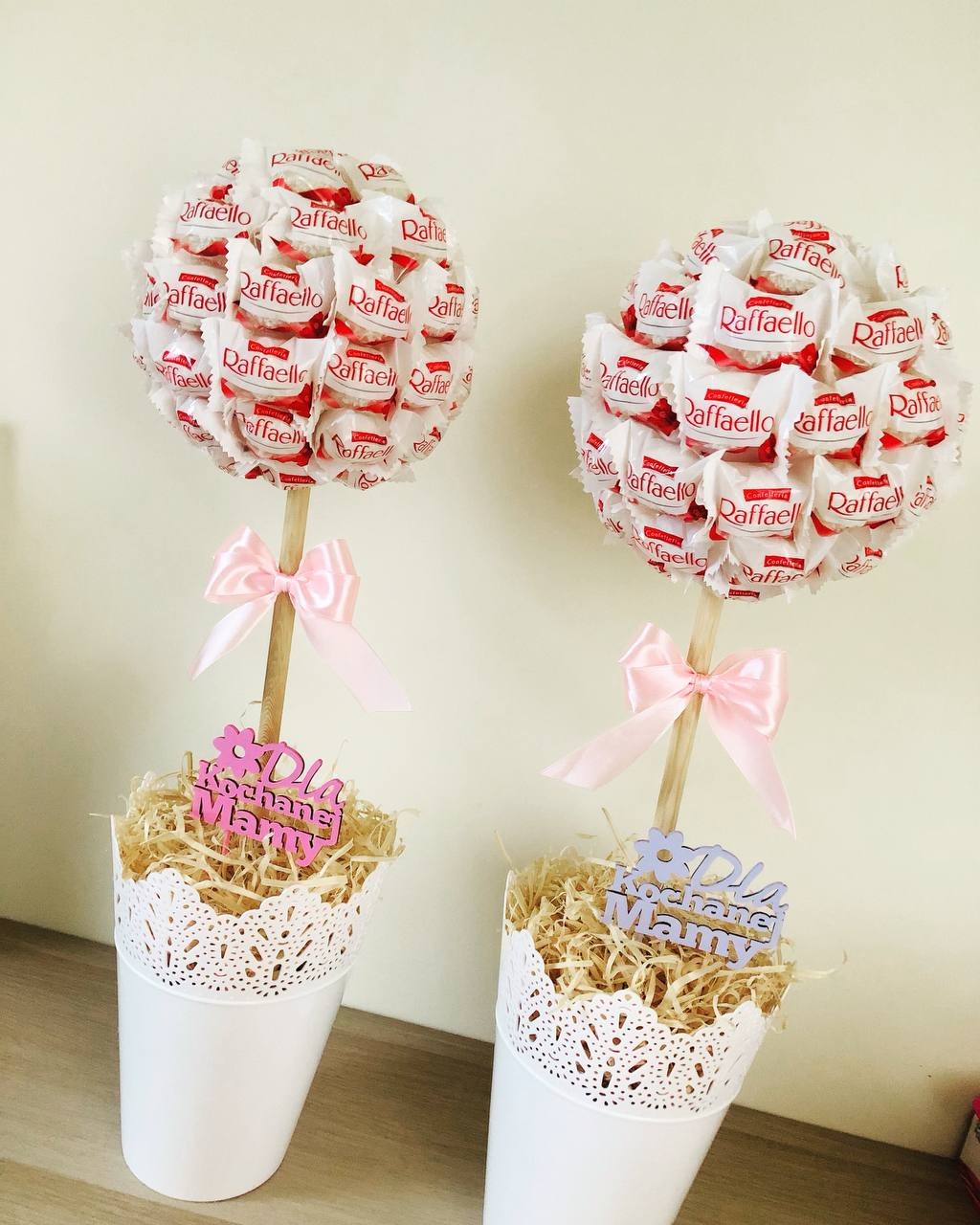 Dwa drzewka z cukierków Raffaello w białych doniczkach z dekoracją koronkową, ozdobione różowymi kokardkami i drewnianymi napisami 'Dla Kochanej Mamy' w kolorach różowym i fioletowym, umieszczone...