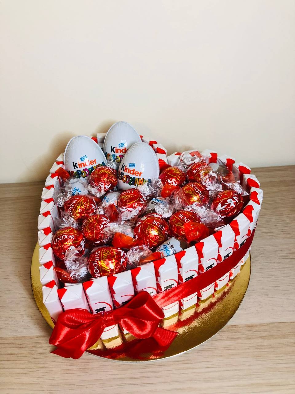 Kompozycja w kształcie serca z czekoladek Lindor, jajek Kinder Niespodzianka, obłożona batonikami Kinder Bueno Mini, przewiązana czerwoną wstążką z kokardą, na złotej podkładce.