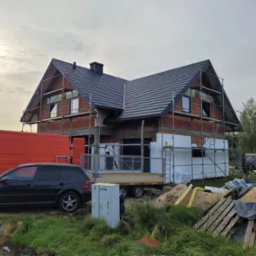 Dom jednorodzinny w trakcie budowy, z widocznym rusztowaniem, częściowo ocieplony styropianem, z nowym dachem z czarnej dachówki, na pierwszym planie zaparkowany samochód osobowy i dostawczy.