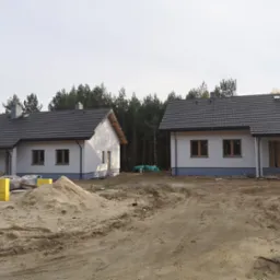 Dwa identyczne, parterowe domy jednorodzinne z szarą dach&oacute;wką, świeżo po budowie, z widocznymi stertami piasku i materiał&oacute;w budowlanych na działce.