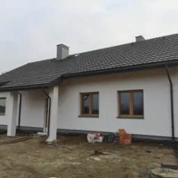 Nowo wybudowany, parterowy dom z białymi ścianami, grafitowym dachem i drewnianymi oknami, w trakcie prac wykończeniowych wokół budynku.