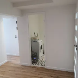 Wnętrze mieszkania po remoncie: świeżo pomalowane białe ściany, drewnopodobna podłoga, widok na łazienkę z pralką i wzorzystą podłogą przez otwarte drzwi.