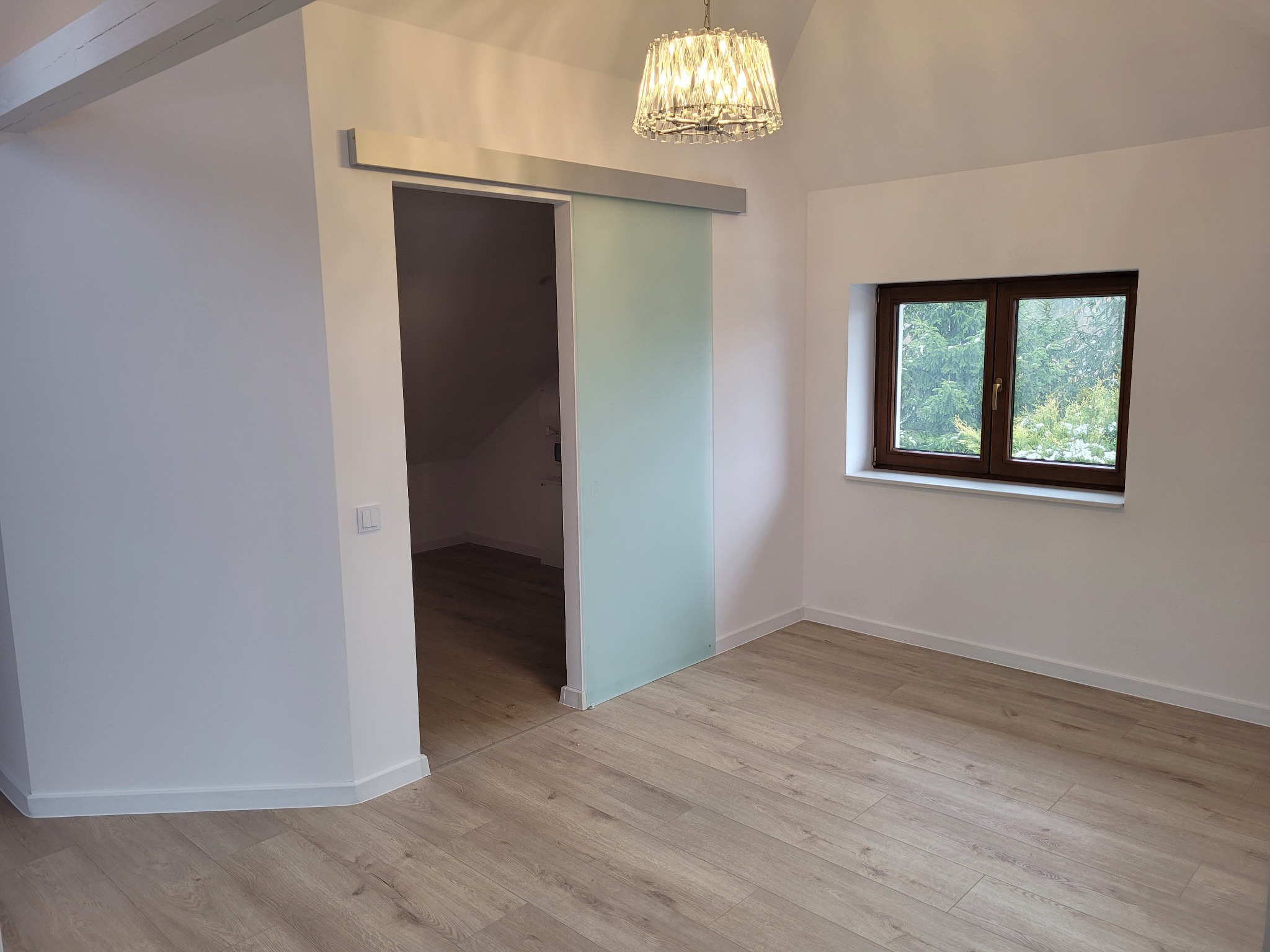 Nowoczesne wnętrze poddasza z przesuwnymi drzwiami ze szkła mlecznego, drewnianą podłogą i oknem z widokiem na zieleń.