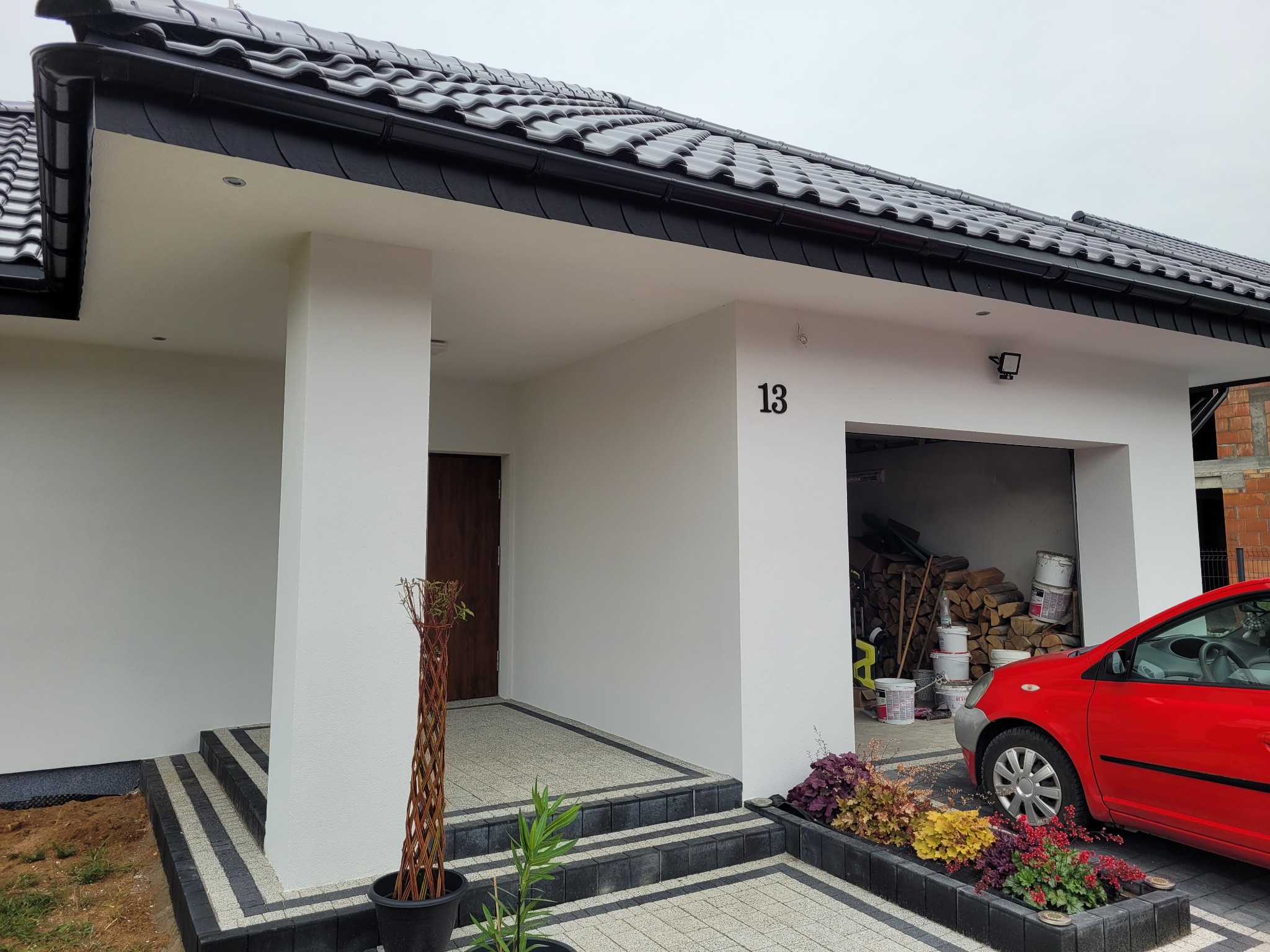 Nowoczesny dom jednorodzinny z białymi ścianami, czarnym dachem i numerem 13 nad garażem, czerwony samochód zaparkowany obok, widoczne elementy wykończenia elewacji.