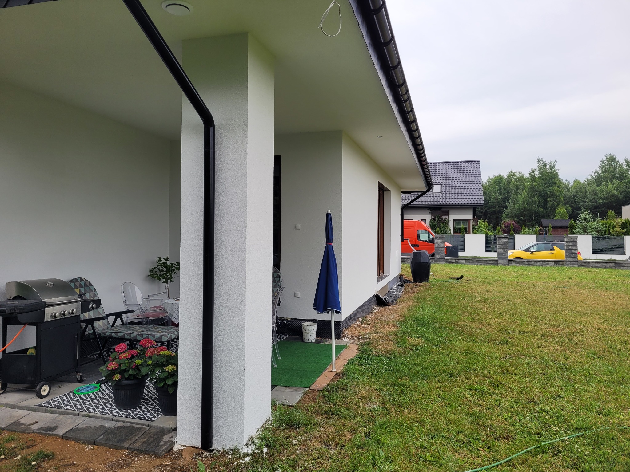 Świeżo otynkowany, biały dom jednorodzinny z czarnymi rynnami, widok z boku na taras z grillem i kwiatami, w tle pomarańczowy samochód dostawczy i żółte auto osobowe za ogrodzeniem.