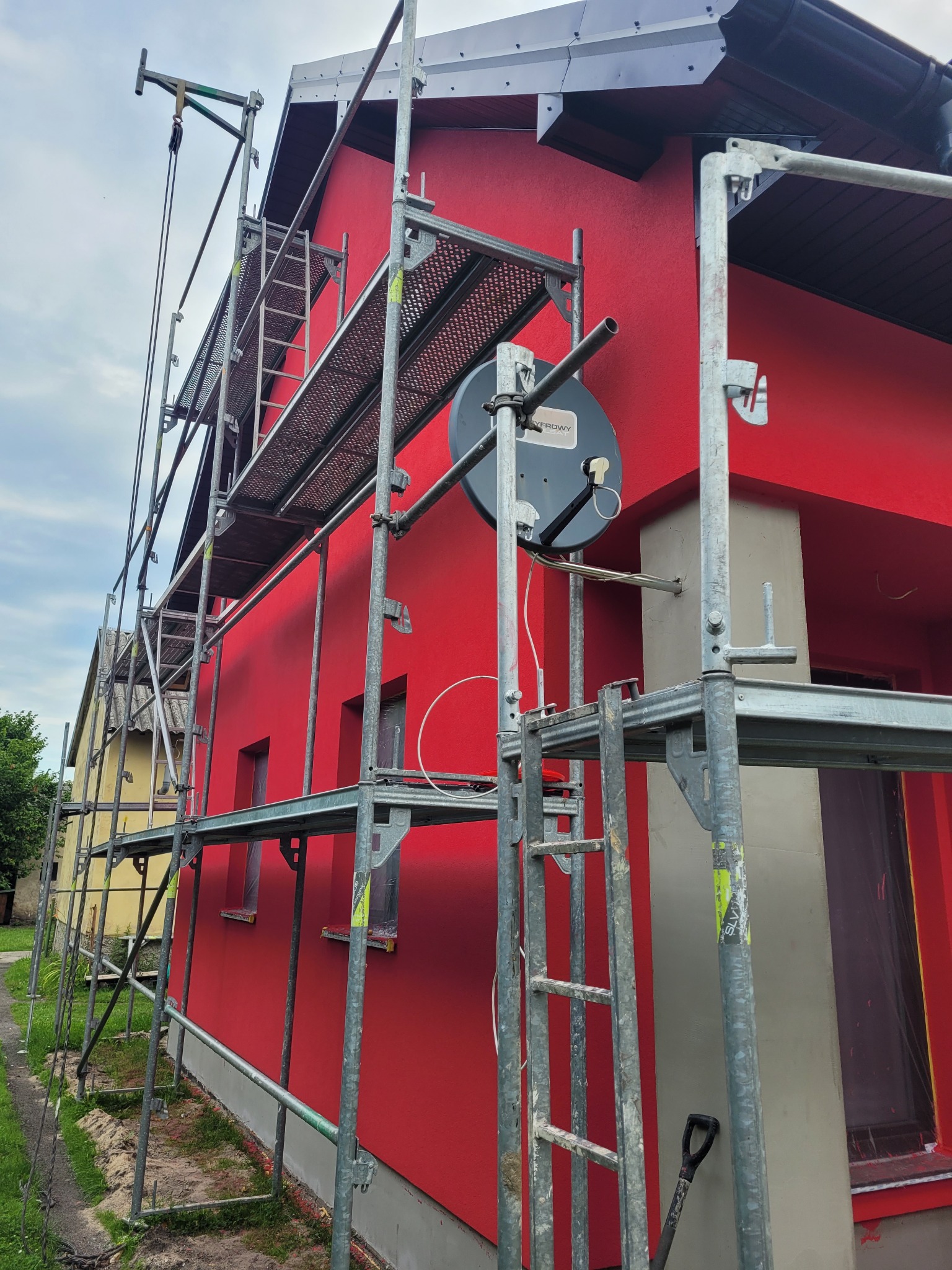 Czerwona elewacja domu w trakcie malowania, widoczne rusztowania, okna zabezpieczone folią, fragment elewacji w kolorze szarym, antena satelitarna, szpadel oparty o rusztowanie.