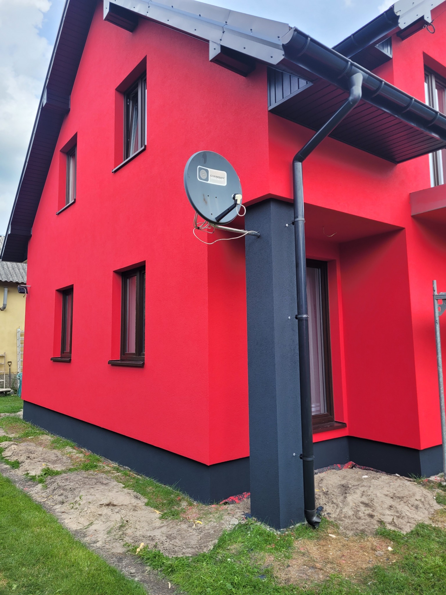 Jaskrawoczerwona elewacja domu jednorodzinnego z ciemnymi oknami i rynnami, widoczna antena satelitarna i fragment trawnika.