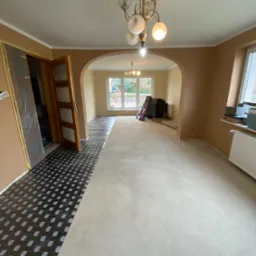 Przygotowanie podłoża pod panele w trakcie remontu mieszkania, widoczna folia izolacyjna na części podłogi oraz wylewka samopoziomująca na pozostałej powierzchni, drzwi wewnętrzne i okna w tle.