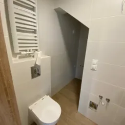 Nowoczesna, biała łazienka z podwieszaną toaletą, grzejnikiem drabinkowym, drewnopodobną podłogą i przygotowanym miejscem na baterię umywalkową.