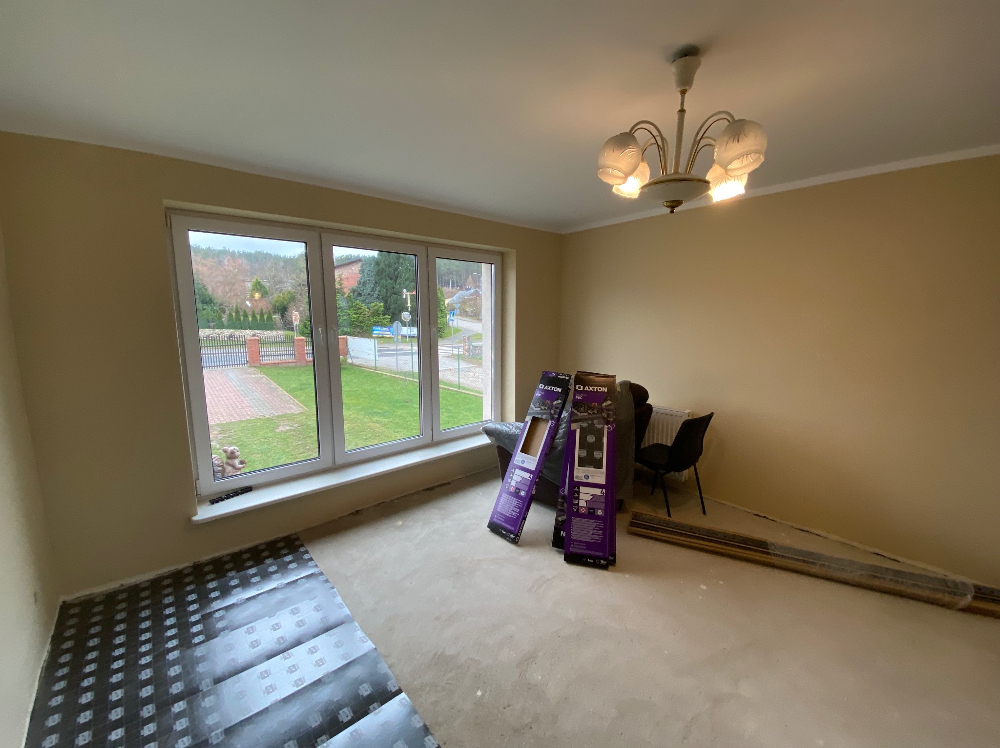 Pokój w trakcie przygotowań do położenia nowej podłogi, widoczne rozłożone podkłady pod panele, paczki paneli Axton oparte o ścianę oraz listwy przypodłogowe w folii.