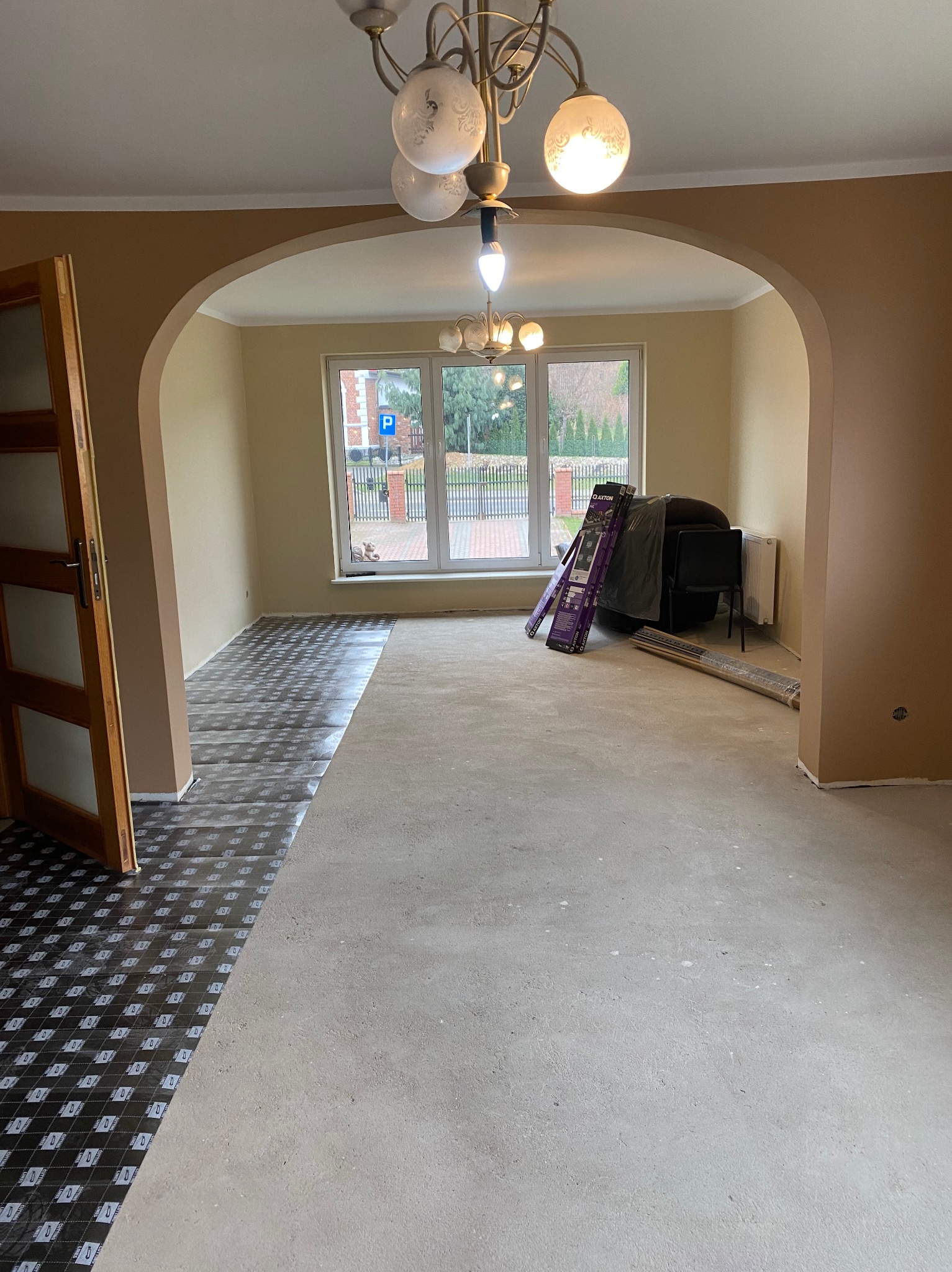 Przygotowanie podłoża pod panele w pokoju z widoczną częścią podkładu i nieprzygotowaną betonową wylewką, widoczne narzędzia i drabina oparta o ścianę przy oknie.