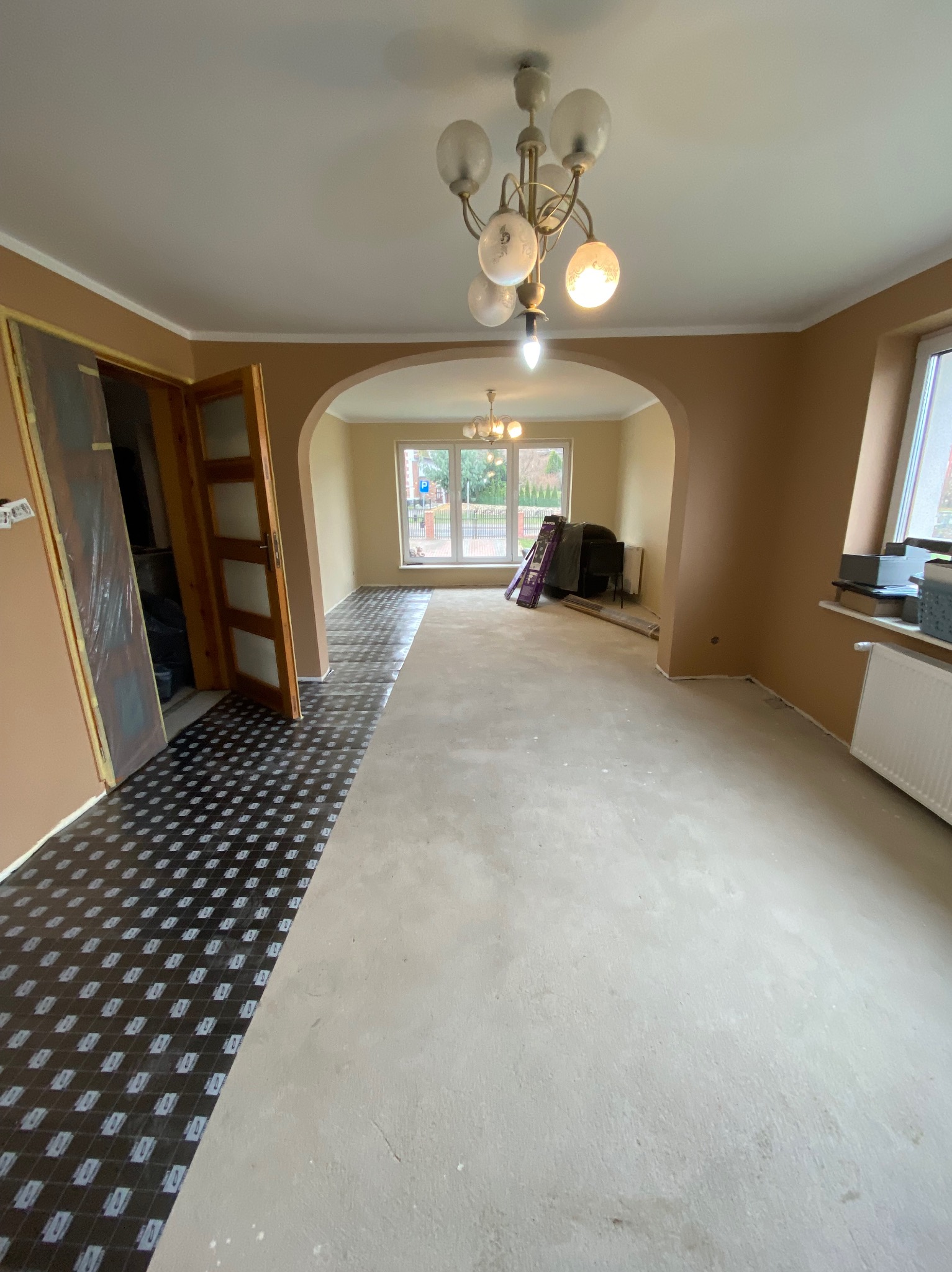 Przygotowanie podłoża pod panele w trakcie remontu mieszkania, widoczna folia izolacyjna na części podłogi oraz wylewka samopoziomująca na pozostałej powierzchni, drzwi wewnętrzne i okna w tle.