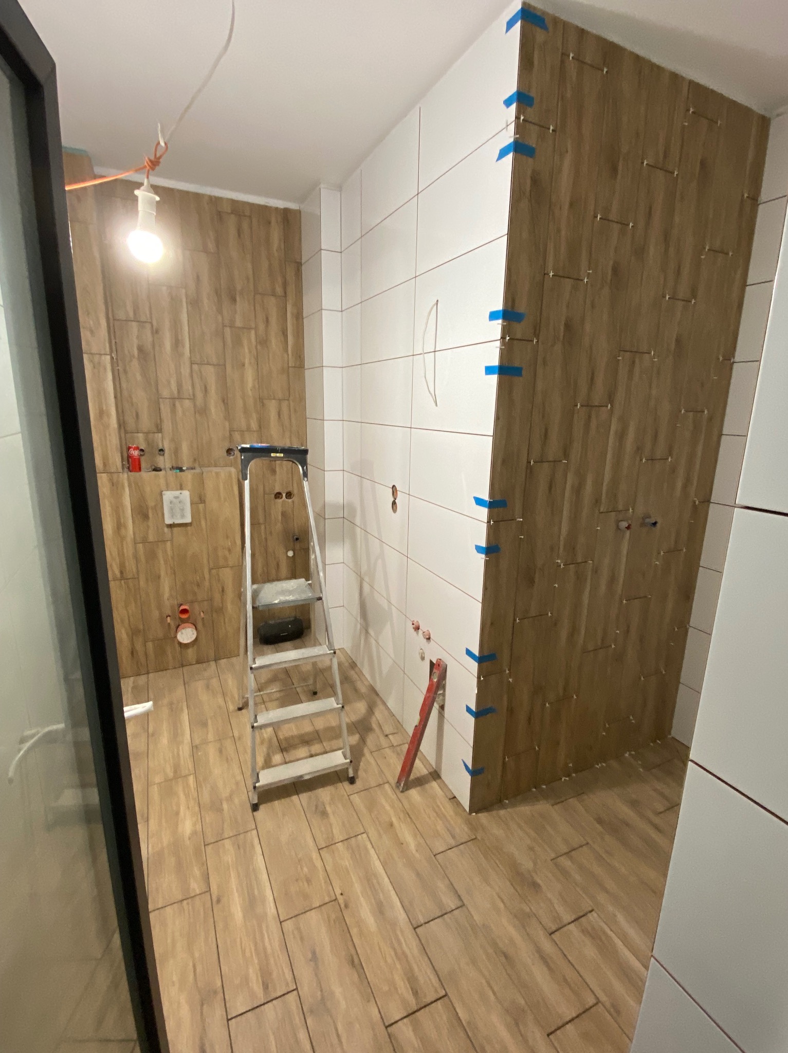 Łazienka w trakcie wykańczania: częściowo ułożone płytki imitujące drewno na ścianie, białe płytki na pozostałych ścianach, drabina w centrum, niebieska taśma zabezpieczająca płytki, widoczne...